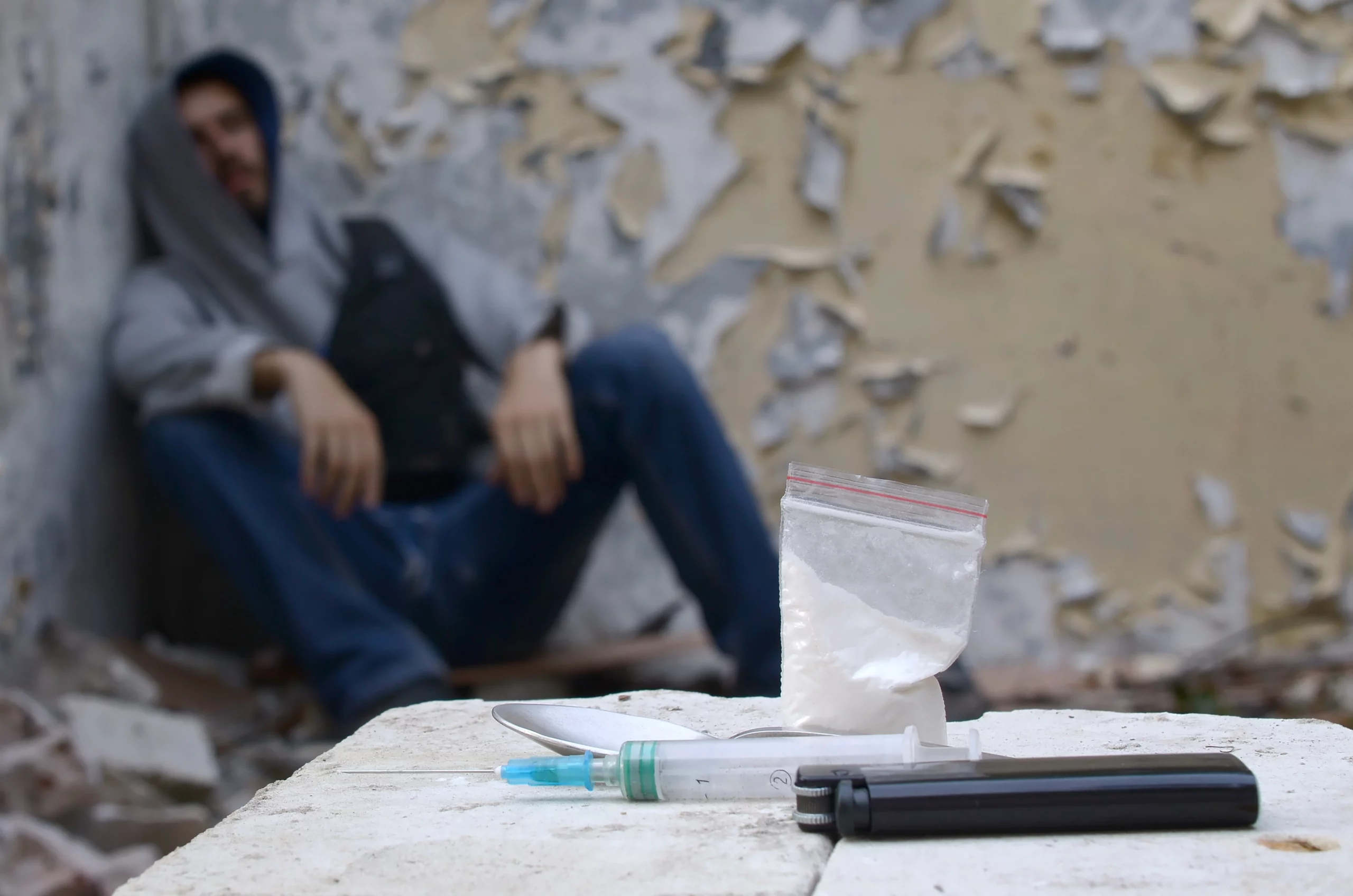 In the foreground, a syringe, spoon, lighter, and baggie of white powder rest on a ledge; a blurred figure sits against a deteriorating wall, suggesting drug use.